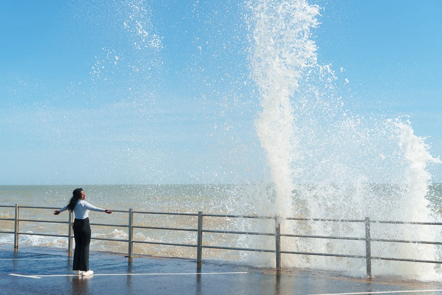 A person standing beside a railing as a wave breaks over the promenade