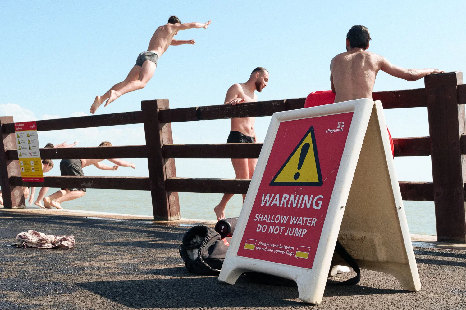 Swimmers jumping from a railing beside a sign warning against jumping