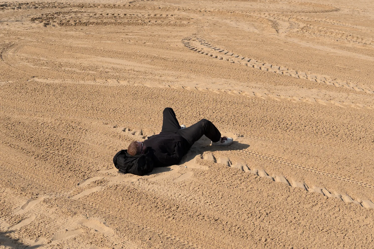 A person lying alone on a wide expanse of sand marked by tracks