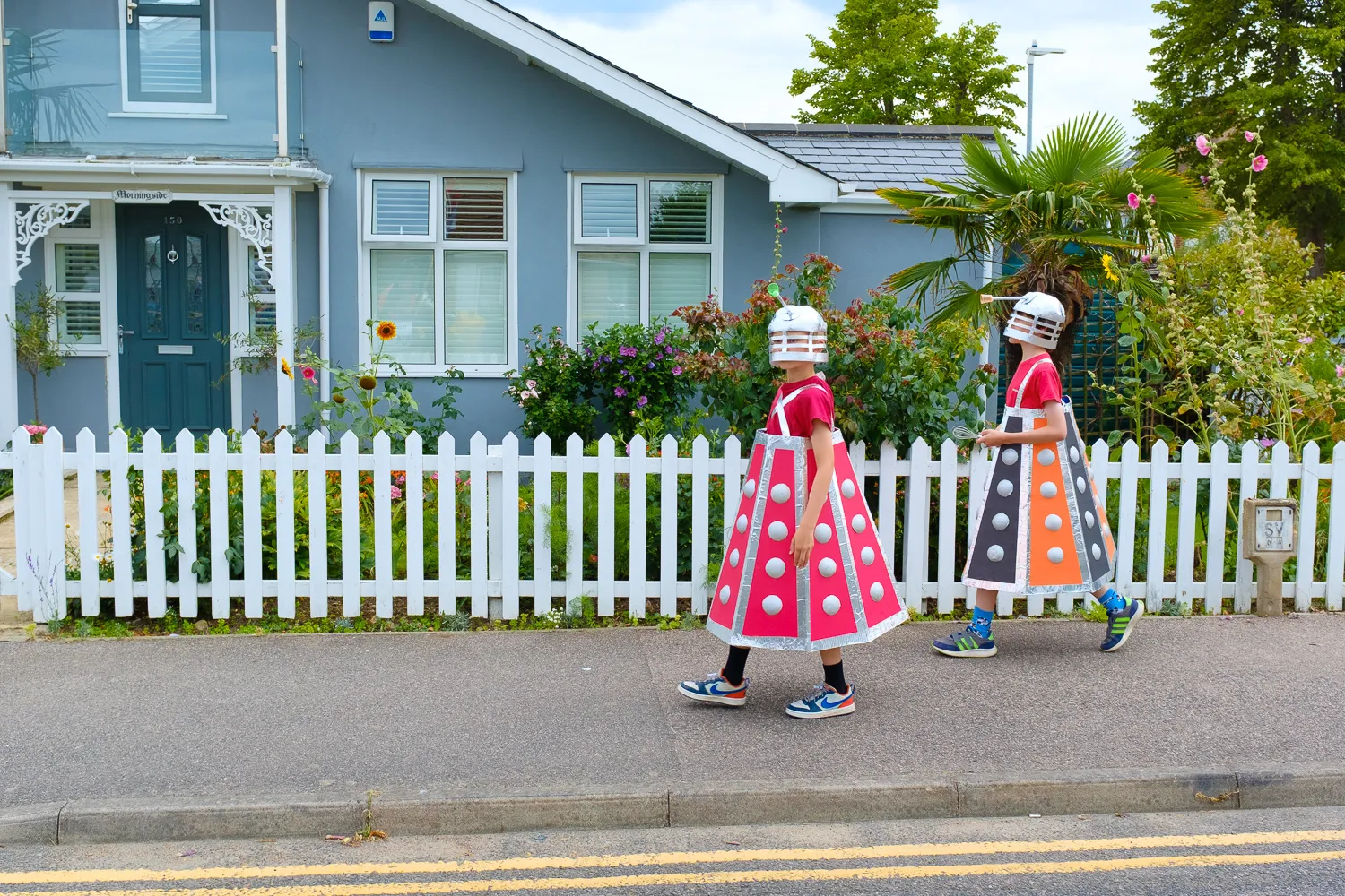 Two people in colourful Dalek costumes walking past a white picket fence